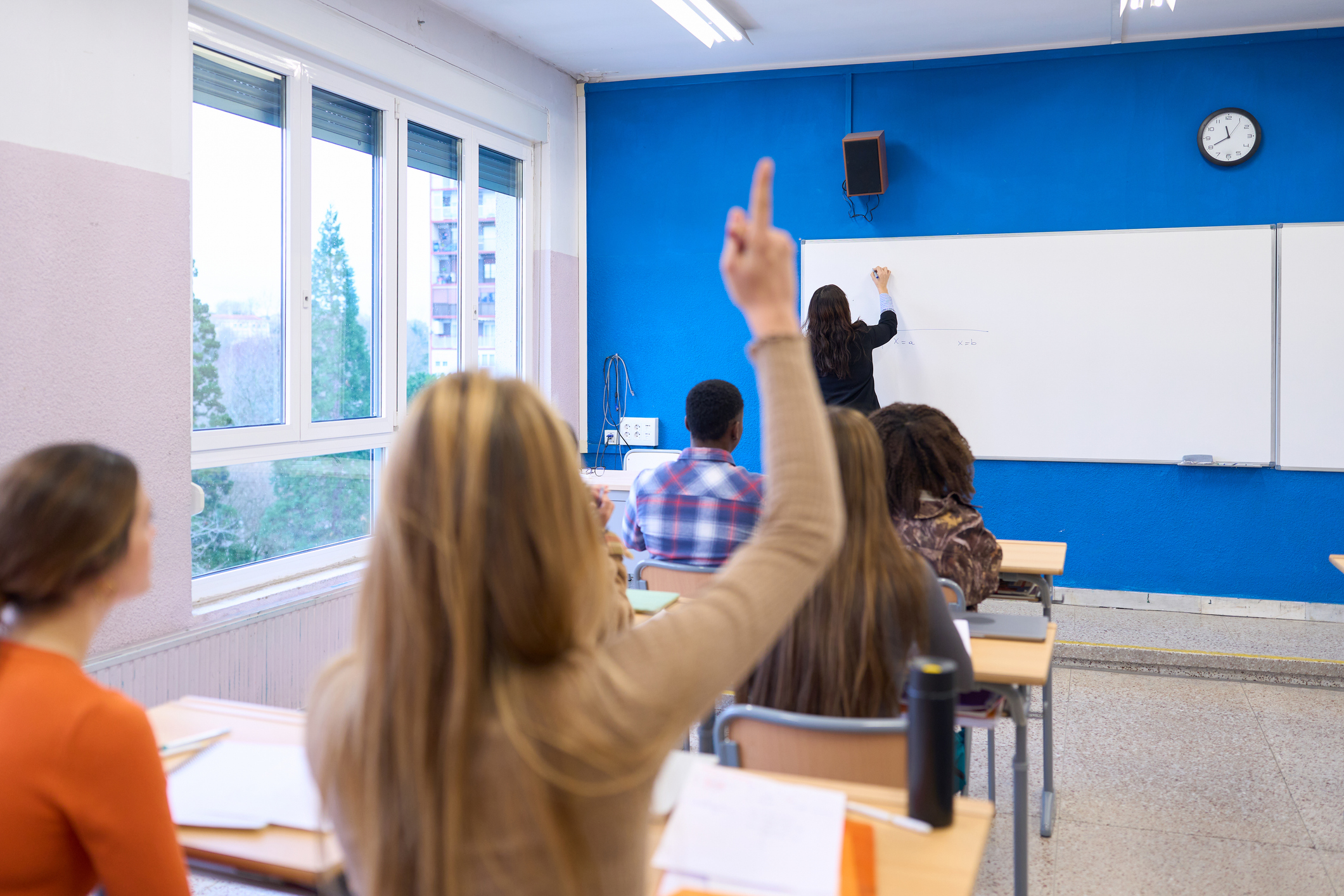 Una professora escriu a la pissarra mentre una alumna aixeca la mà. 