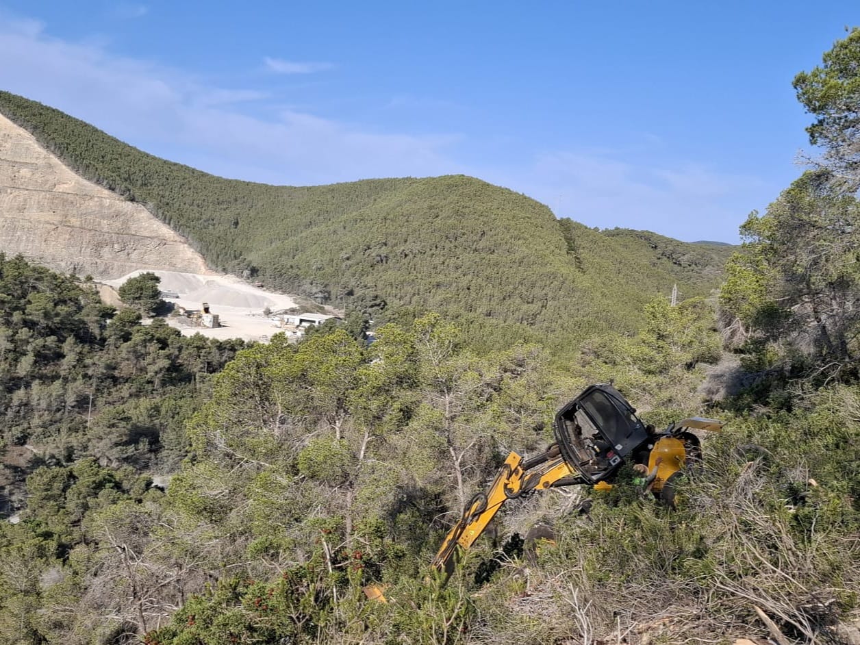 Una màquina treballa entre la vegetació d’una zona forestal, en una àrea de muntanya.